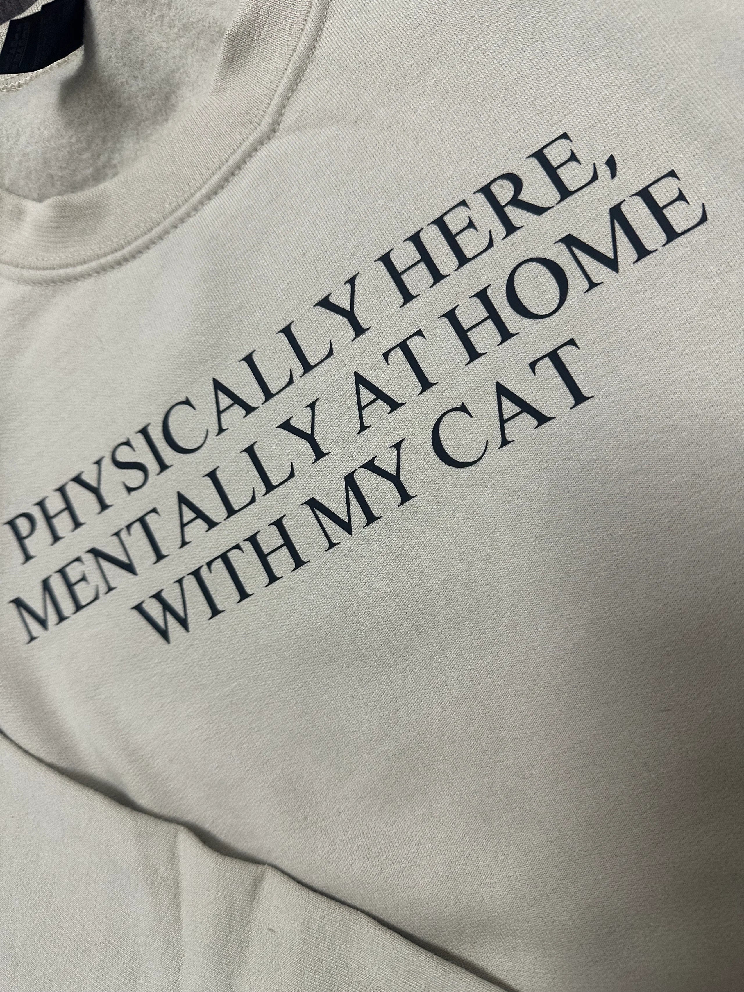 Beige sweatshirt with text 'Physically here, mentally at home with my cat' on a light background